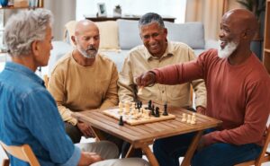 A group of seniors hanging out around a table, as two of them play chest against each other. One senior elder is moving a piece on the chessboard