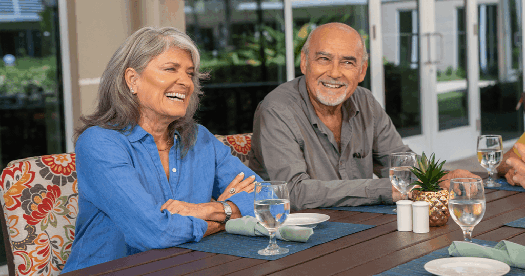 A cheerful couple enjoys a meal outdoors with glasses and plates on a wooden table.