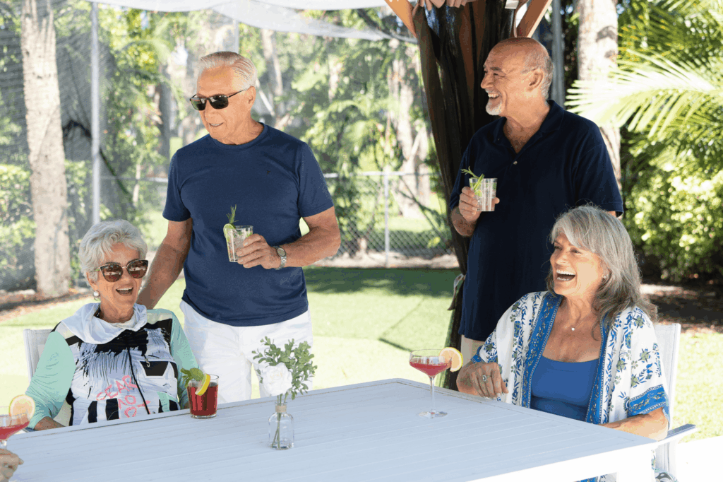 Four older adults enjoy drinks and laughter at an outdoor table setting with greenery behind them.