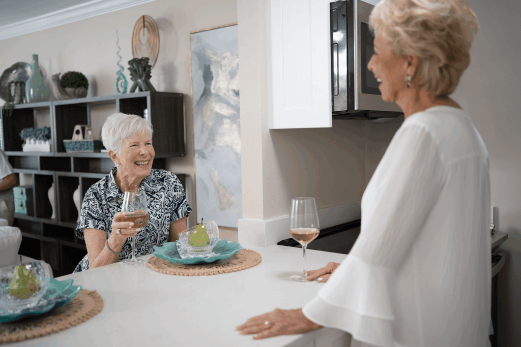 Two women share laughter and wine at a kitchen counter with decorative items around them.