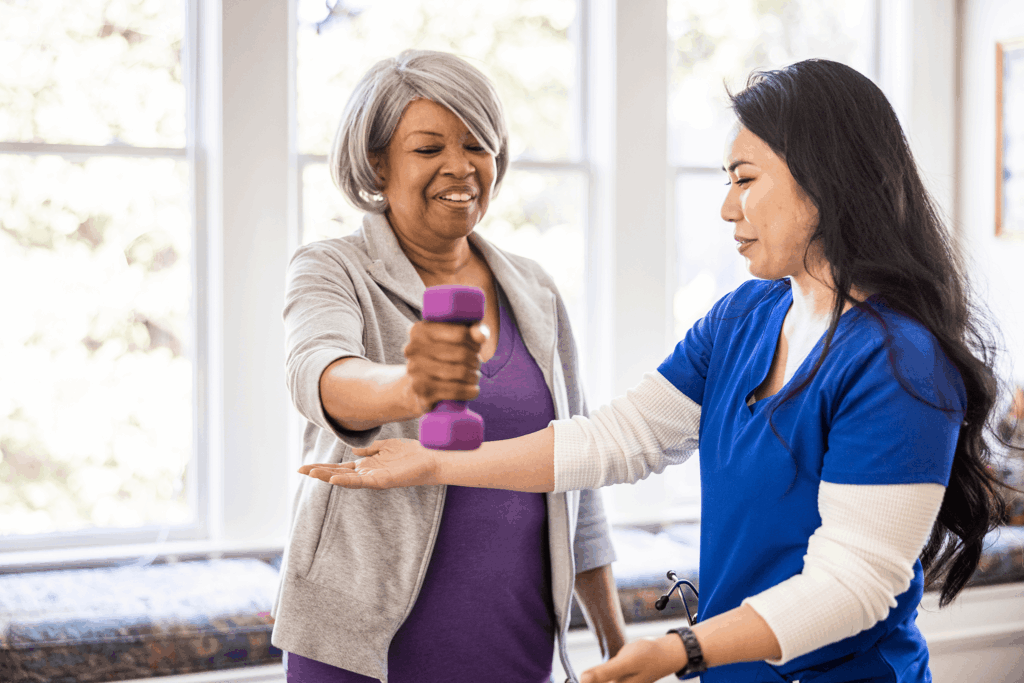A caregiver assists an older woman with arm exercises using weights indoors near windows.