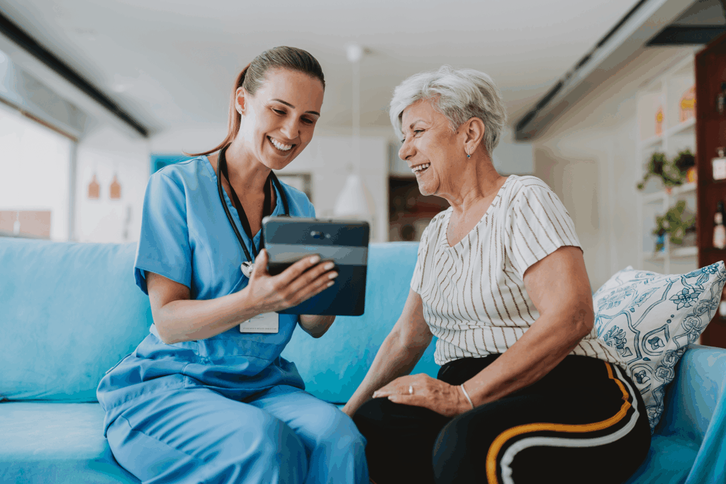 Nurse and elderly woman share smile on blue couch with tablet.