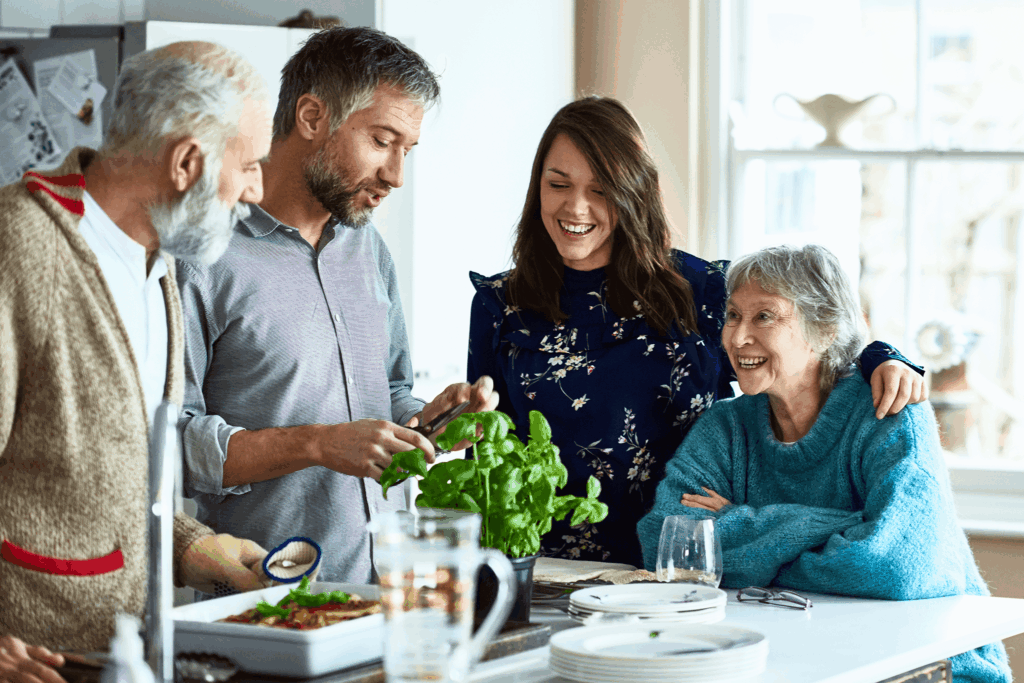 A group gathers around a table with food and greenery, sharing laughter and conversation.