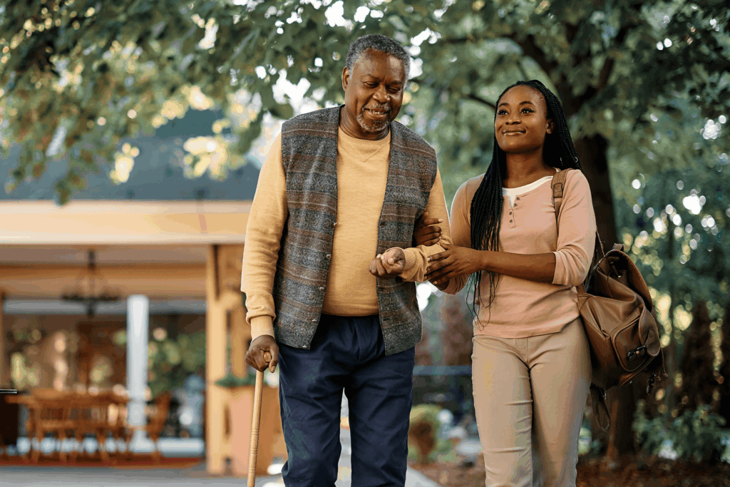 A smiling older man and younger woman walk together outdoors under trees.