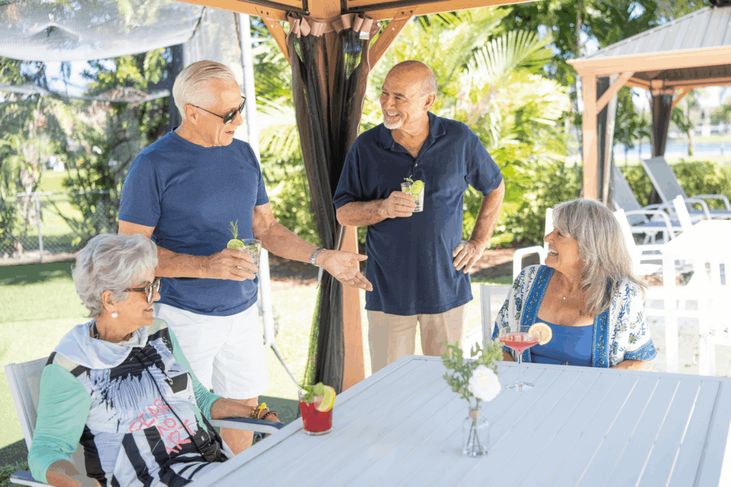Four seniors enjoy drinks and conversation at an outdoor table under shade.