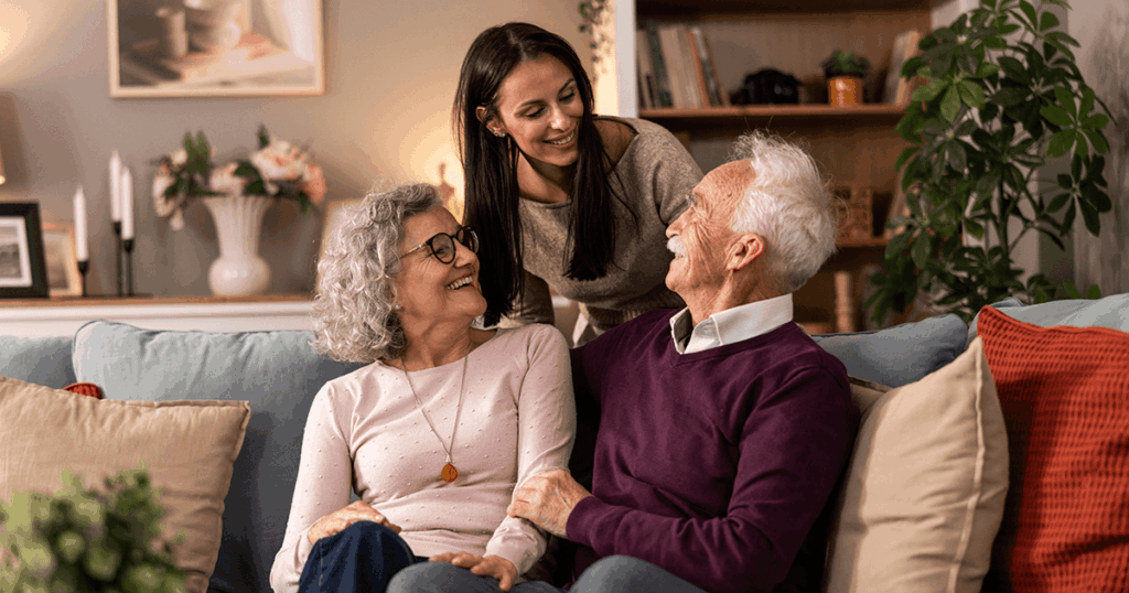 Three people share laughter on a cozy couch with warm decor and plants around them.