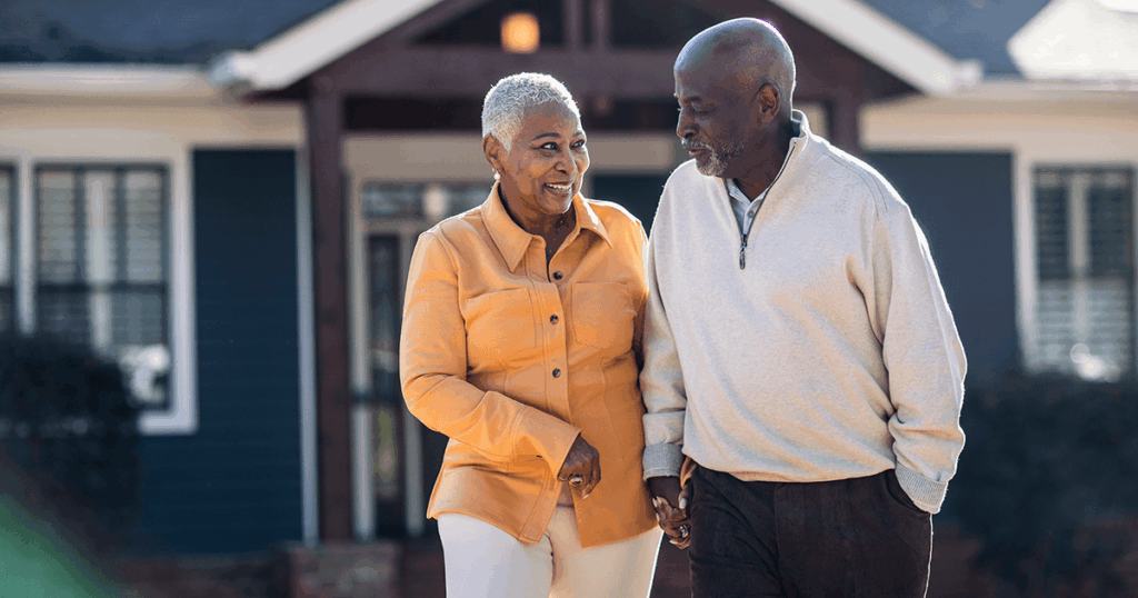 A happy elderly couple walks hand-in-hand outside their home on a sunny day.