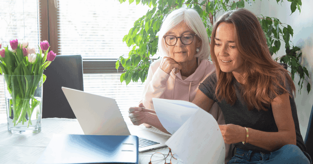 Two women review documents and laptop together at table with flowers nearby.