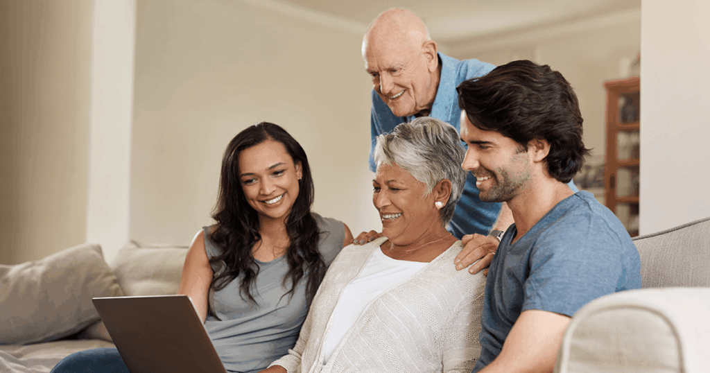 A family gathers around a laptop on a couch, smiling and laughing together.