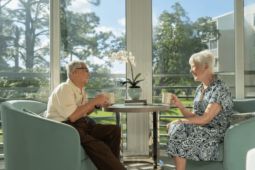 Elderly couple enjoys coffee on balcony with scenic view and potted orchid.