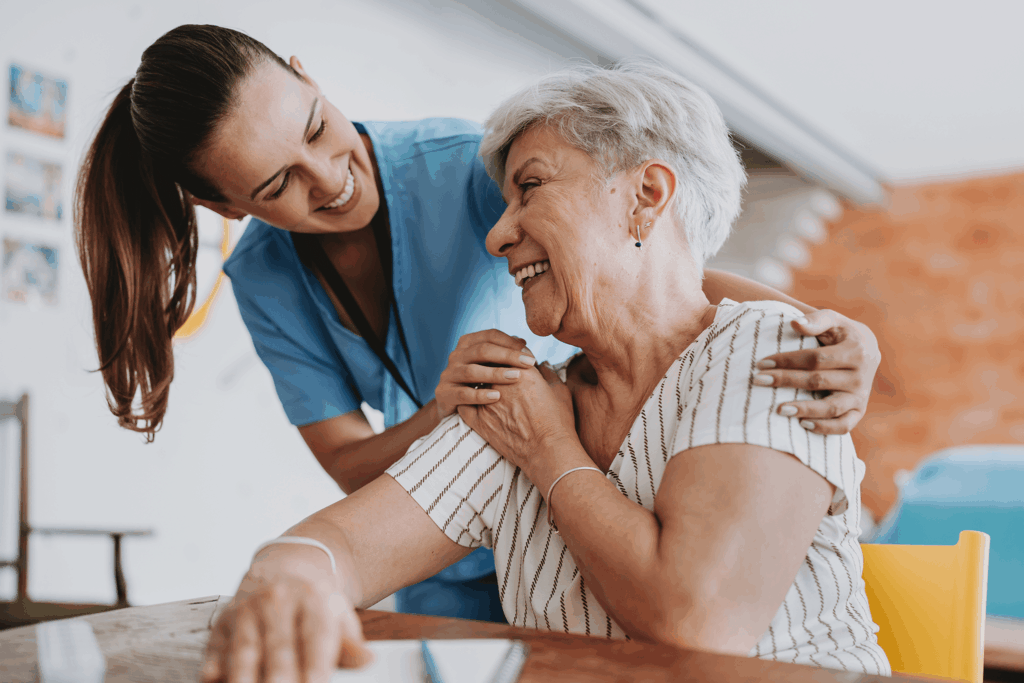 A caregiver smiles as they assist an elderly woman at a table indoors.