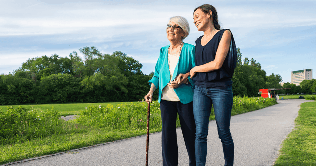Two women walk together on a path surrounded by greenery under a blue sky.