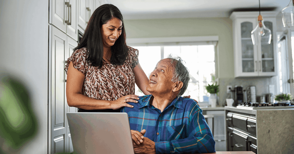 A woman assists an older man using a laptop in a kitchen setting.