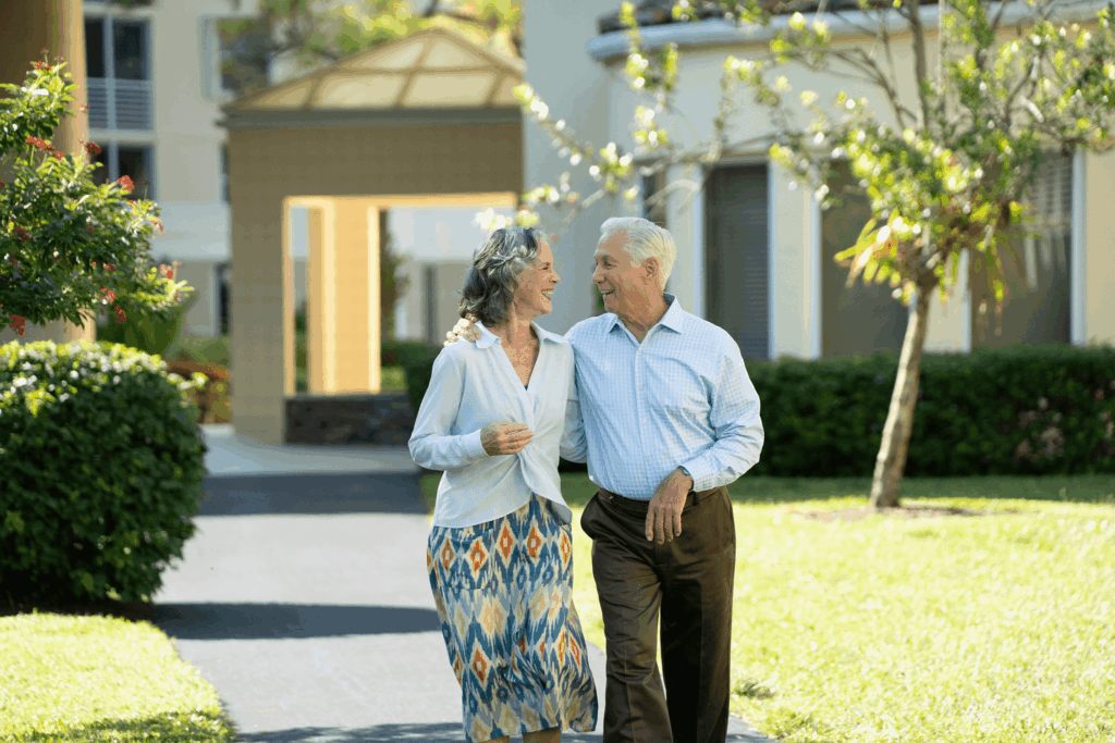 A happy couple walks hand-in-hand through a sunny garden path near a building with an arched entrance and greenery around