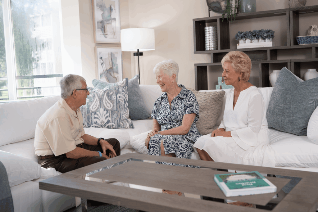 Three people sit on white couches talking and laughing in a cozy living room with modern decor.