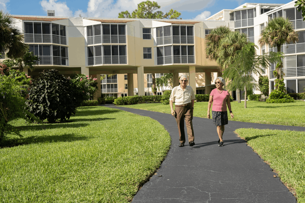 A couple walks along a paved path surrounded by lush greenery and palm trees near an apartment complex on a sunny day.