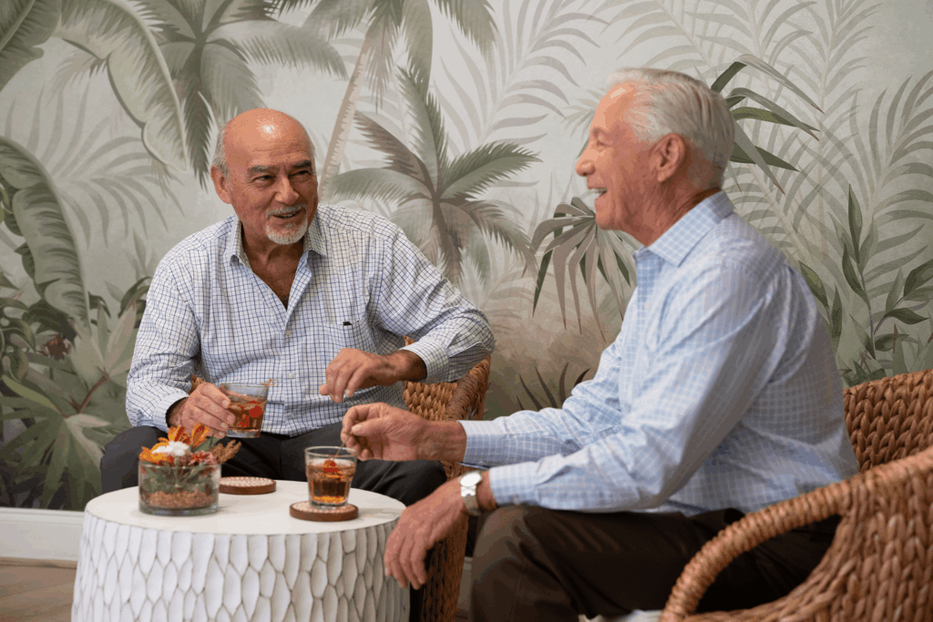Two men sit at a small table with drinks and snacks, smiling and talking against tropical leaf wallpaper.