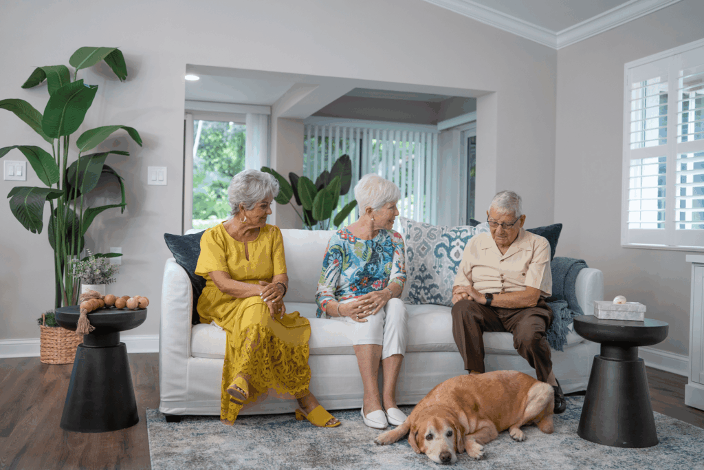 Three elderly individuals sit on a white couch with a dog resting nearby in a cozy living room setting.