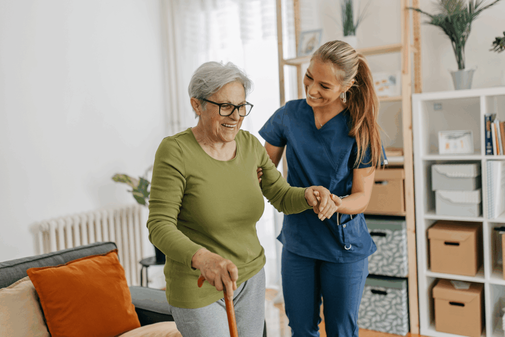 A caregiver assists an elderly woman with walking using a cane indoors.