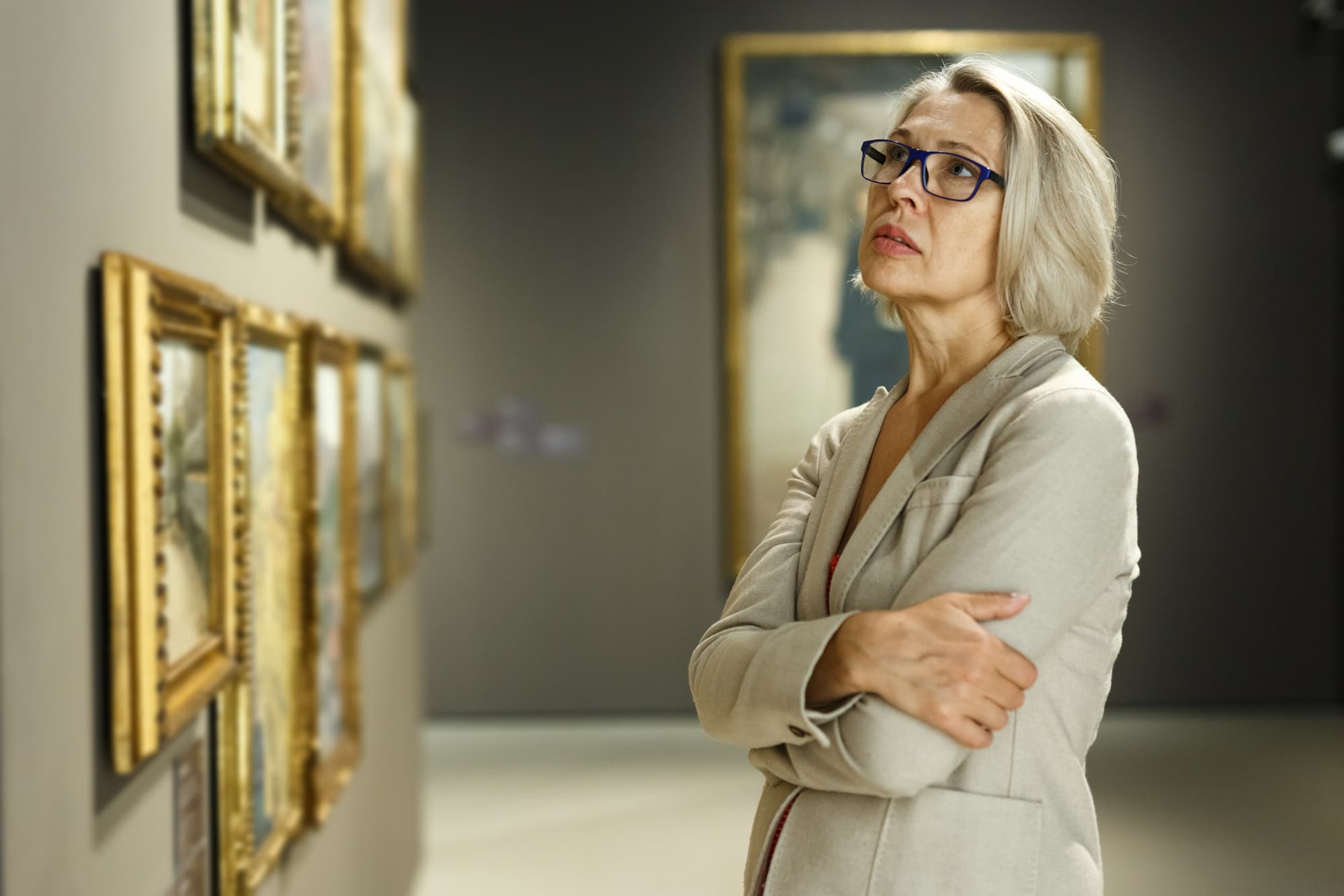 A senior woman dressed in white with her arms crossed staring at an art piece pensively in thought