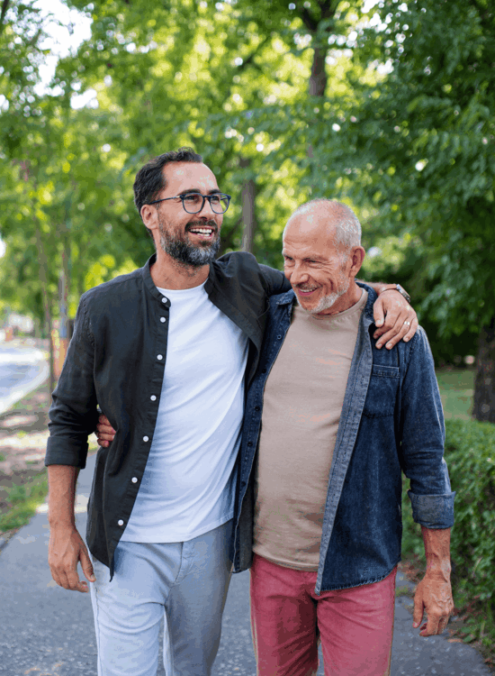Two men walk arm-in-arm on a sunny path surrounded by greenery.