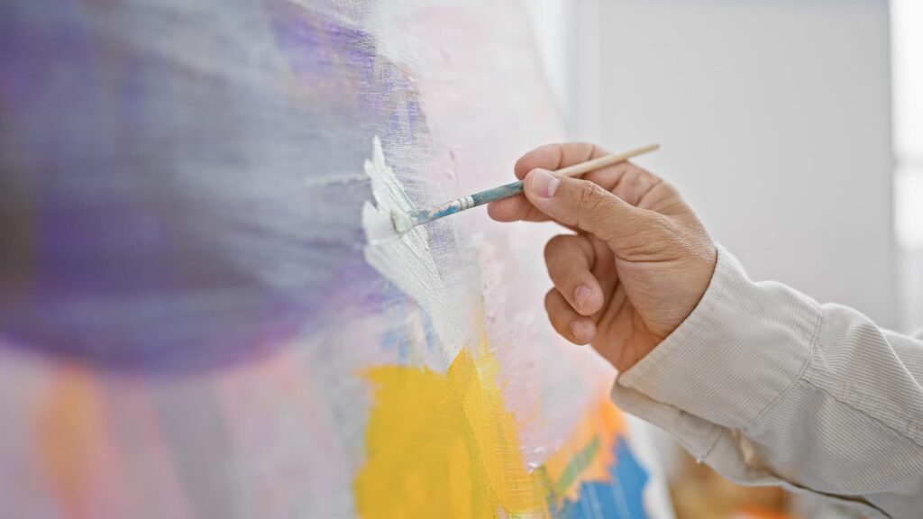 a hand holding a brush as it paints white onto a canvas full of colors