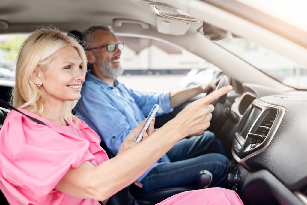 An elderly man driving a car as an elderly woman navigates him from the passenger seat with her phone