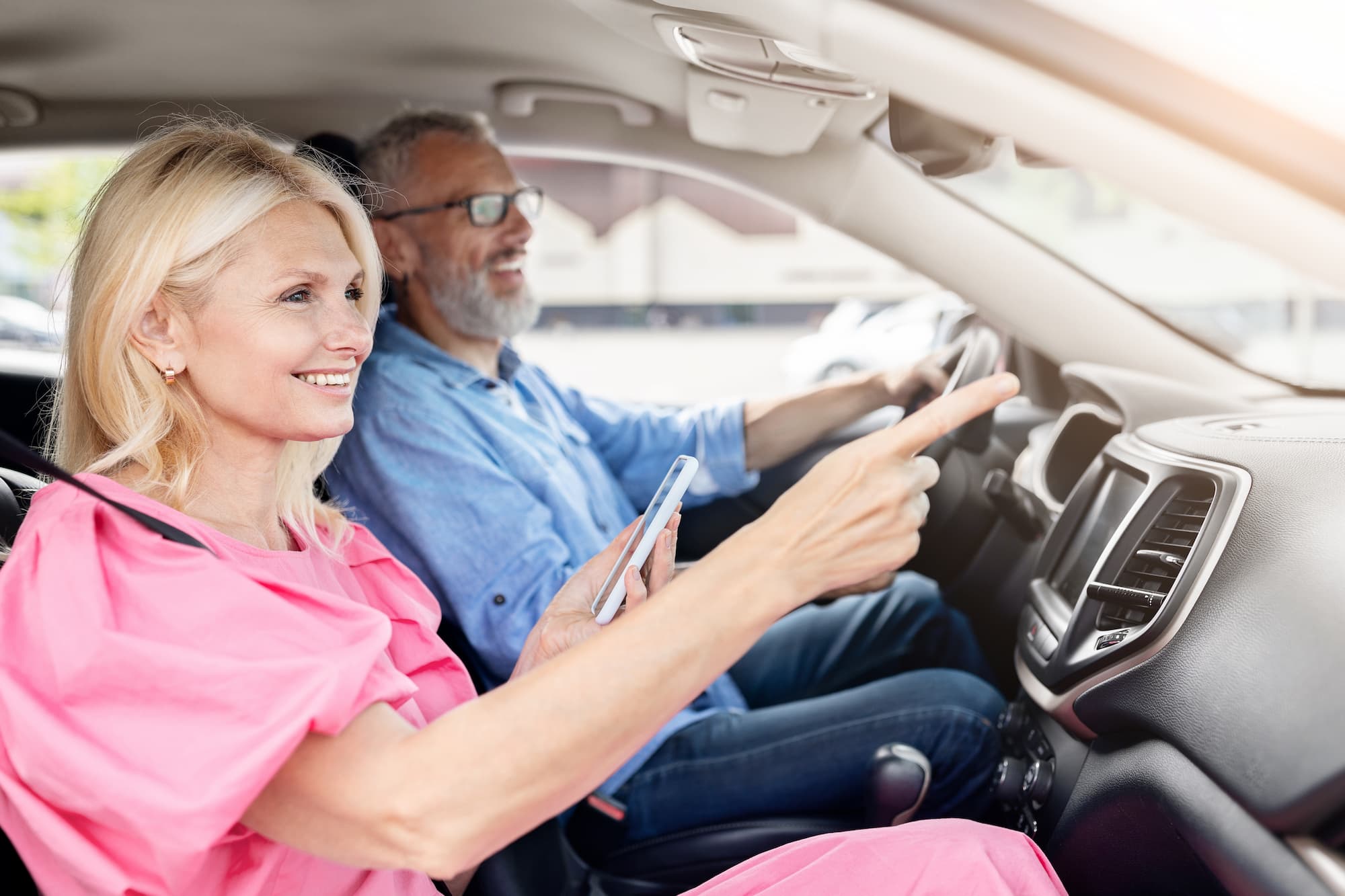 An elderly man driving a car as an elderly woman navigates him from the passenger seat with her phone